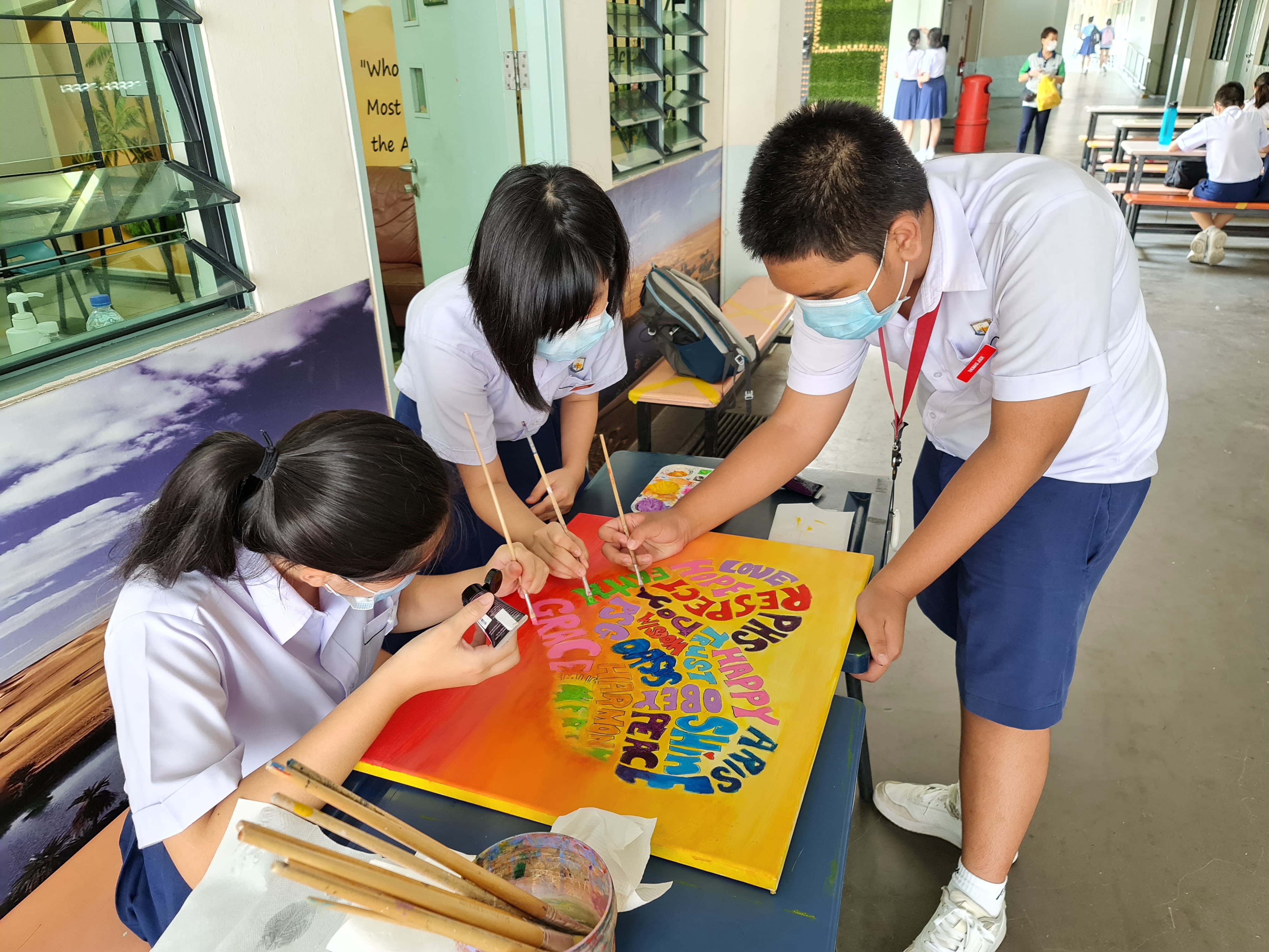 Three masked school students in uniforms paint colorful lettered words on a large canvas at a table.